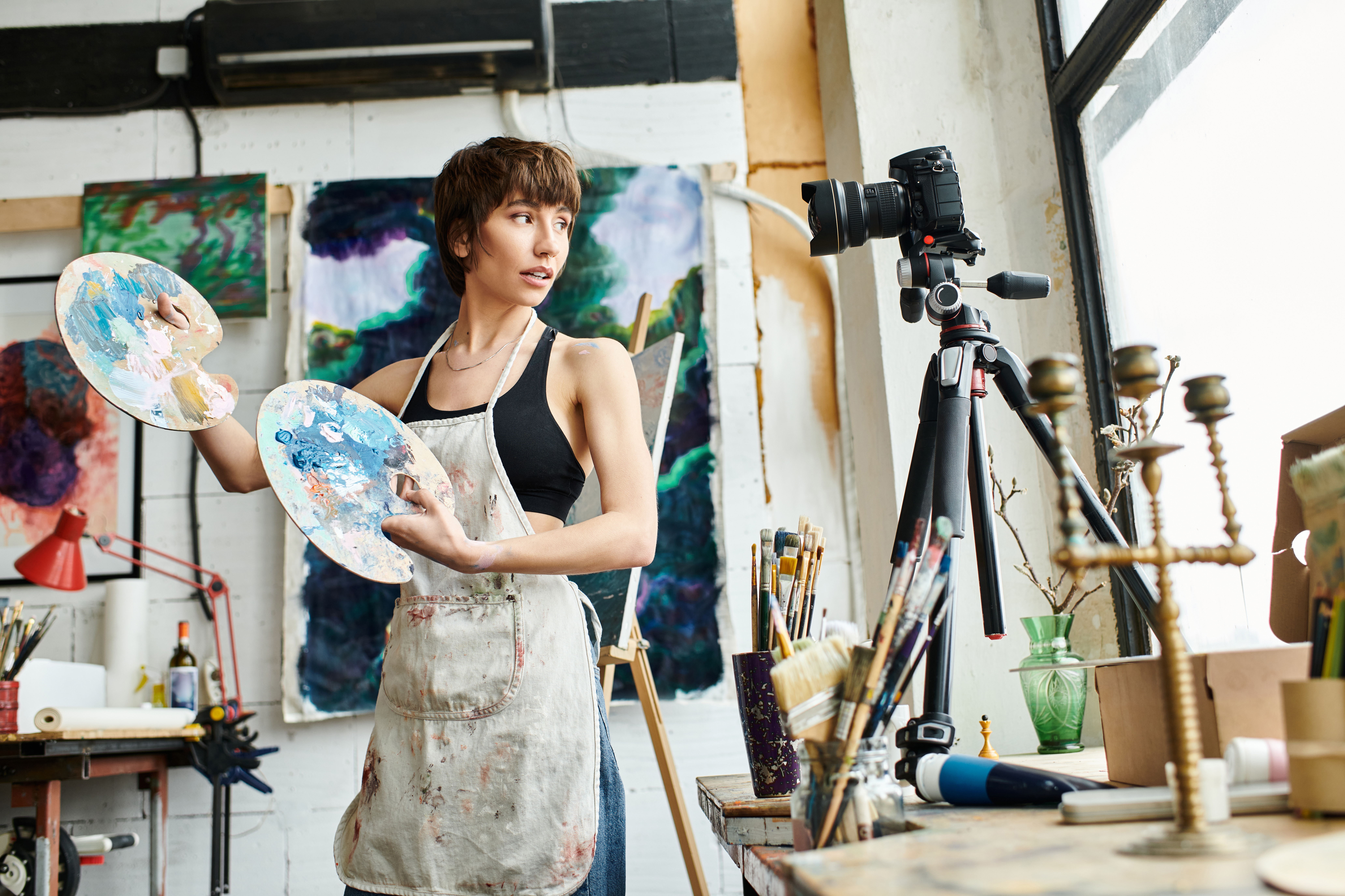 A female artist holds palettes toward a camera on a tripod in a well lit painting studio setting.