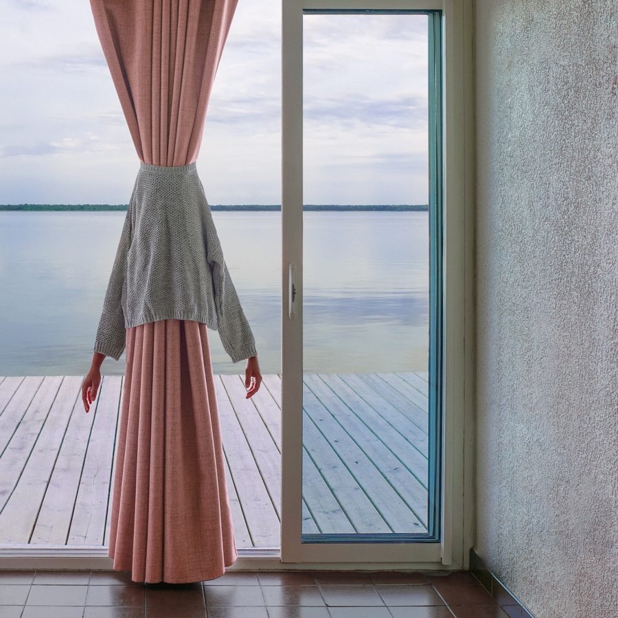 view from inside a room with glass slideing doors, looking out at a deck over glassy water. A gray sweater is around a long pink curtain, a woman's hands emerging from the sleeves of the sweater. Image by Émilie Möri