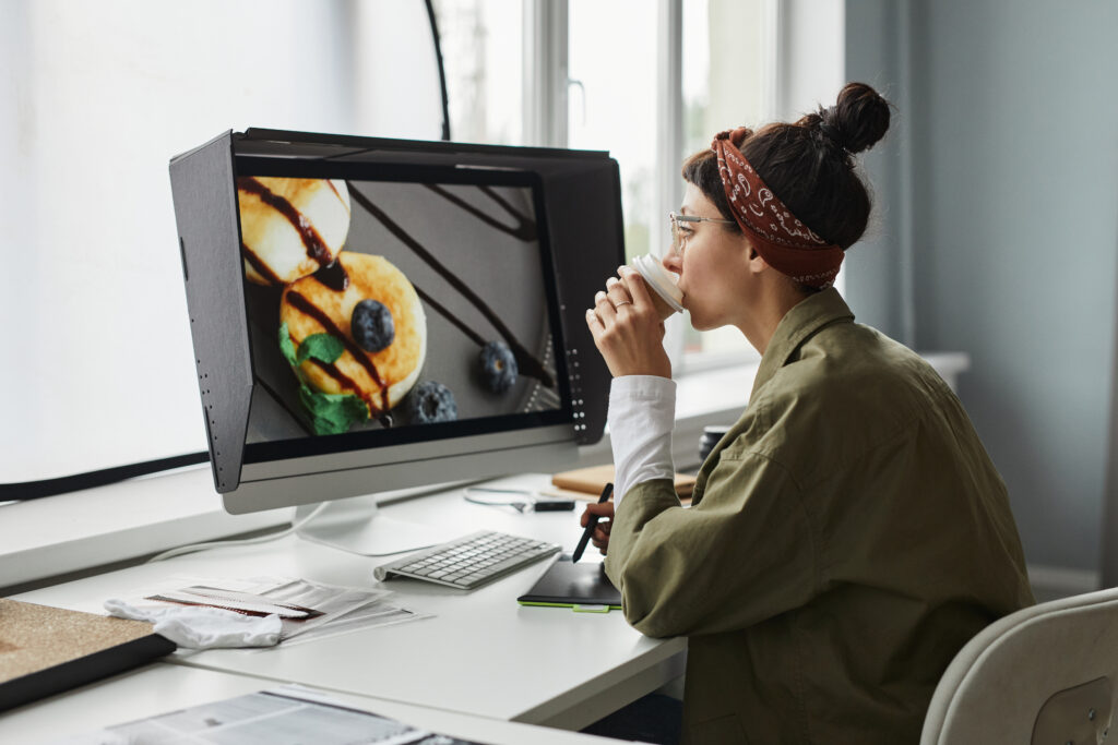 female photographer editing food photos on desktop computer while drinking coffe