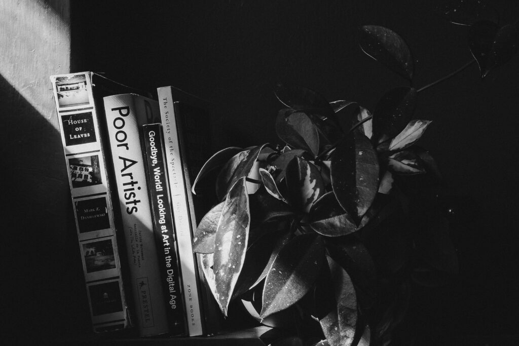 black and white image of a bookshelf holding books beside a plant: "house of leaves", "poor artists", "Goodbye, World!", "the society of the spectacle"