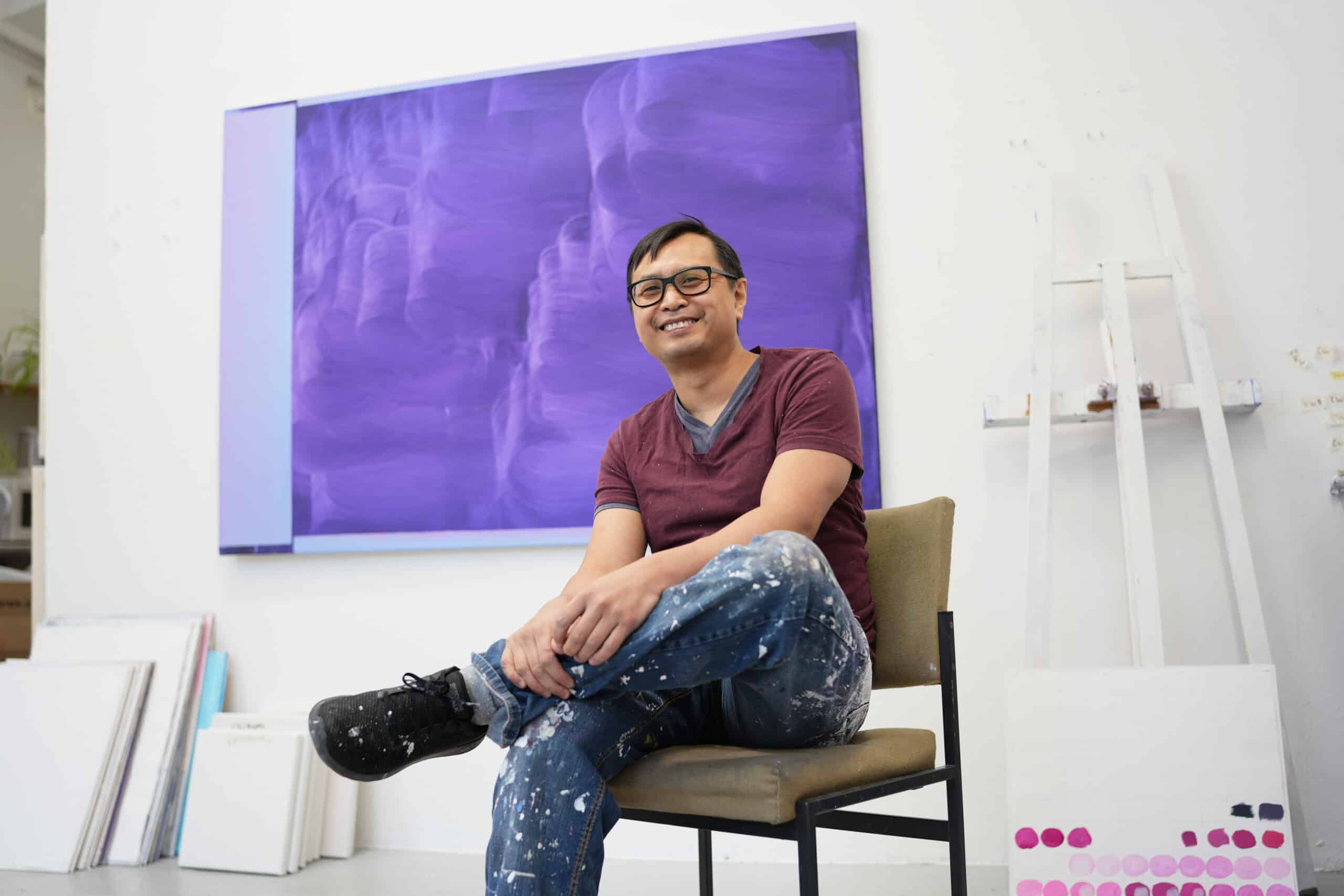 Portrait of Nam Duc Nguyen smiling and looking at the camera, sitting in a chair in front of a purple painting with large brush marks in his studio.